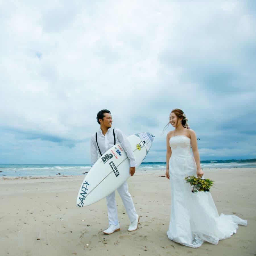 海岸にて新郎新婦二人歩いている新郎はサーフボードを持って