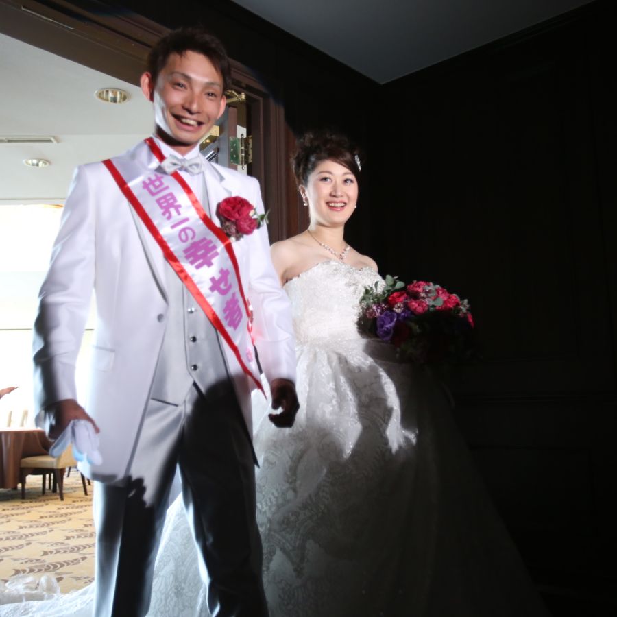 世界一の幸せ者のタスキをかけた新郎と白ドレスの新婦が披露宴の入場